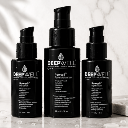 Three DEEPWELL skincare bottles on a reflective marble surface with a marble wall background. The center bottle is a black black-violet pump bottle labeled 'Powered Face Moisturizer', flanked by two black pump bottles labeled 'Powered Day Serum' and 'Powered Night Serum'.